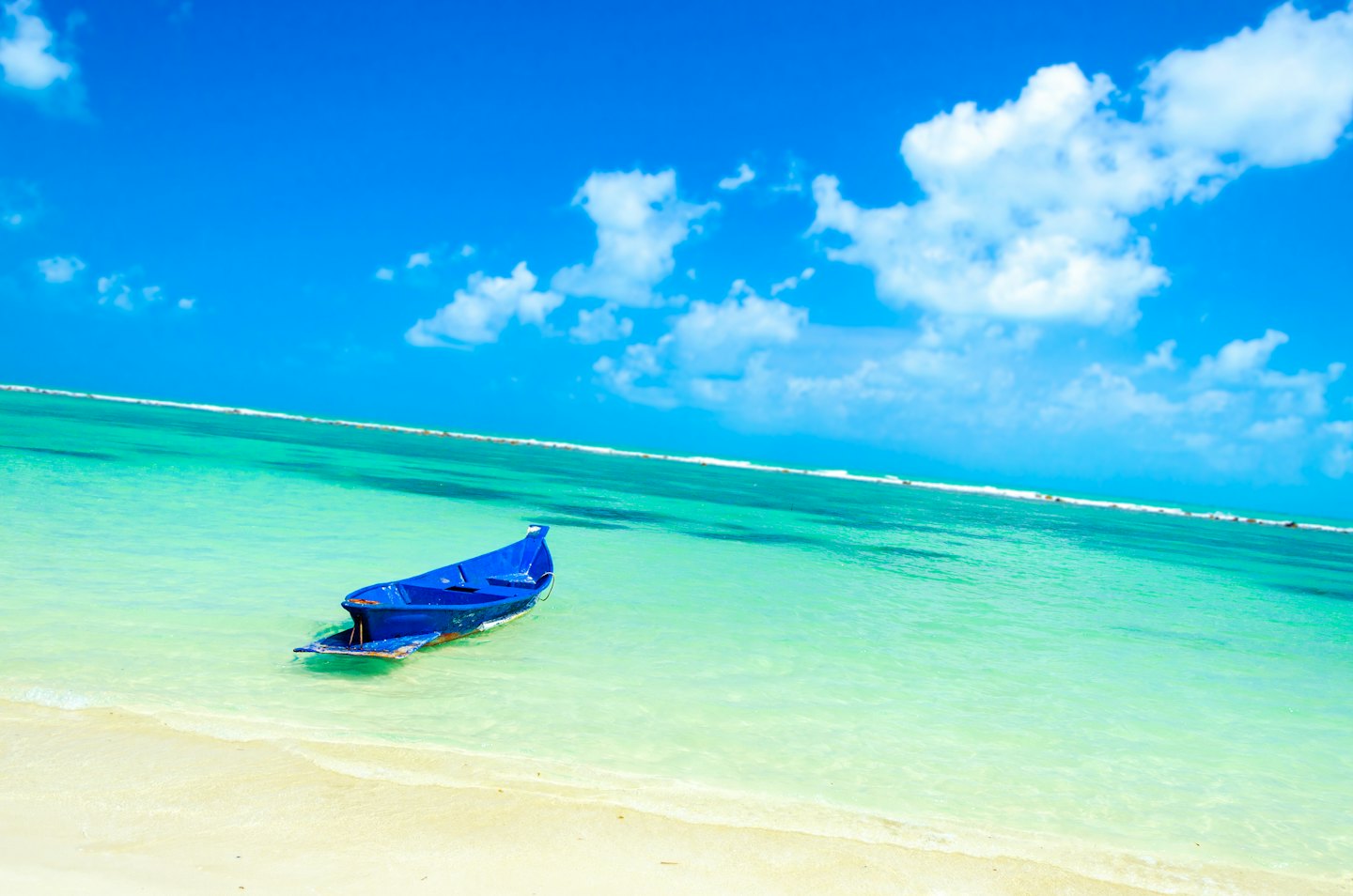 Un bote azul descansa cerca de una playa de arena en aguas poco profundas de color turquesa.